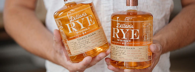Two bottles of Dillon's Rye Whiskey being held by a man with a white button down shirt.