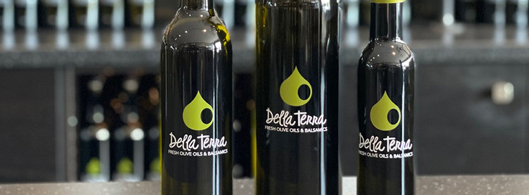 Three bottles of Della Terra premium extra virgin olive oil in glass bottles decorated with applied ceramic labelling printing process by Stanpac.